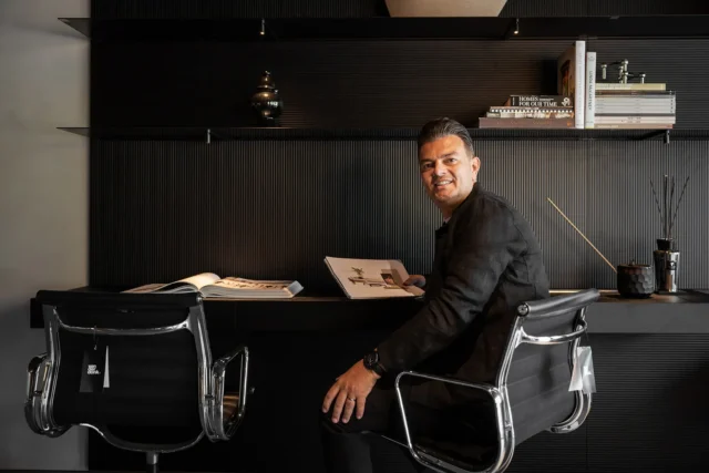 A photo of a man sitting at a desk. He is turned to the camera and holding a brochure.